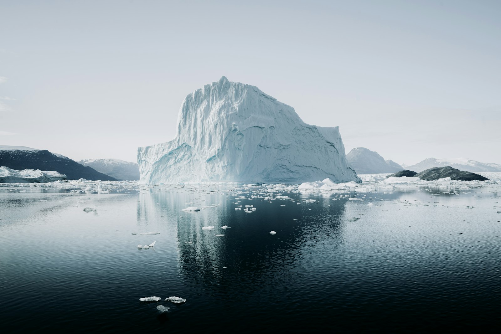 A towering Greenland iceberg mirror-reflected on glassy still water with distant snow-capped peaks